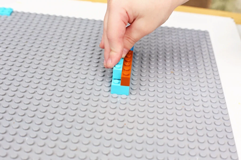 Hand placing blue and orange LEGO bricks on a large gray baseplate (close-up)​