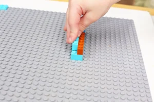 Hand placing blue and orange LEGO bricks on a large gray baseplate (close-up)​