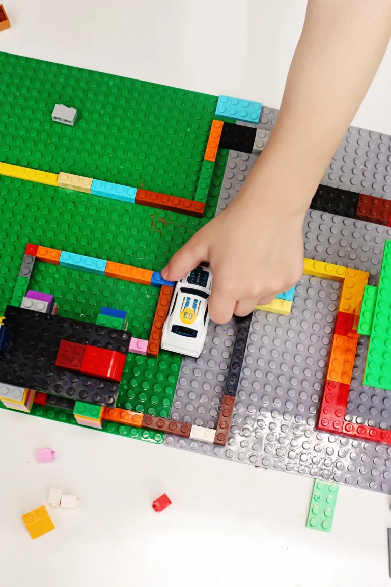 Child's hand places a white toy race car onto a colorful LEGO board on a green base plate.