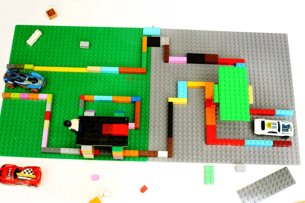 Top-down view of a colorful LEGO build on green and gray baseplates; a blue racing car sits on the left with many bricks around it.