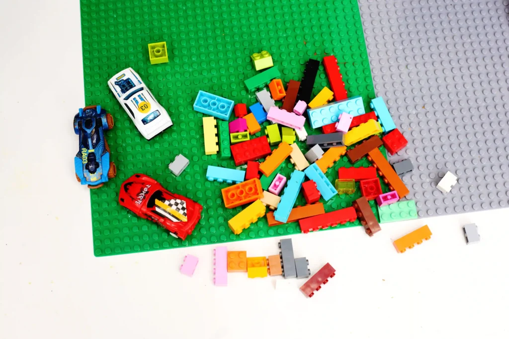 supplies for the LEGO maze for cars: Green LEGO baseplate with a jumble of colorful bricks scattered across, plus two toy race cars on the left (a white race car and a blue race car) and a red vehicle at the bottom left.