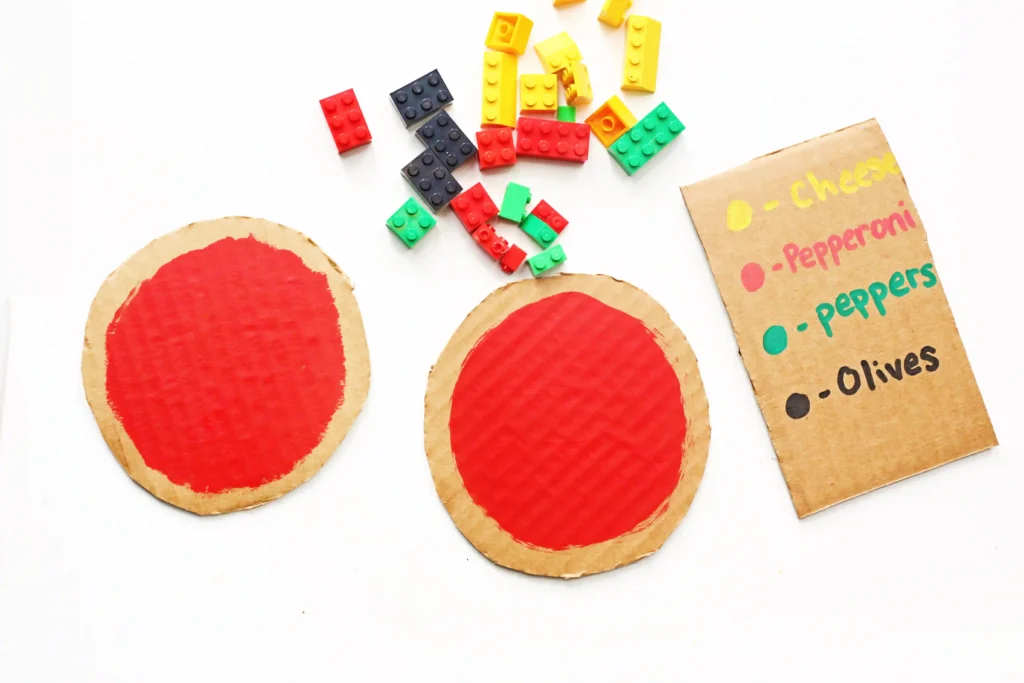 Assorted colorful LEGO bricks scattered on a white surface beside round cardboard pizza templates with red centers and a menu listing toppings like cheese, pepperoni, and olives.