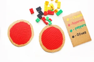 Assorted colorful LEGO bricks scattered on a white surface beside round cardboard pizza templates with red centers and a menu listing toppings like cheese, pepperoni, and olives.