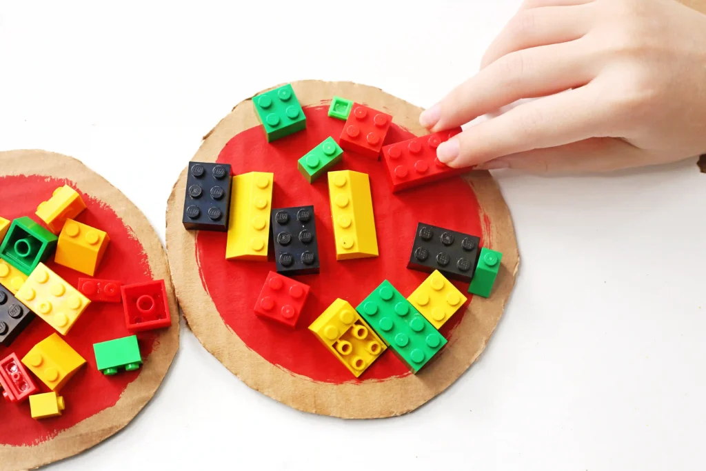 Hand places a red 2x2 LEGO brick onto a red-painted circular cardboard surface with other colorful bricks nearby.