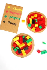 Cardboard pizza with red sauce topped by colorful LEGO bricks; a handwritten list shows toppings: cheese, pepperoni, peppers, olives.