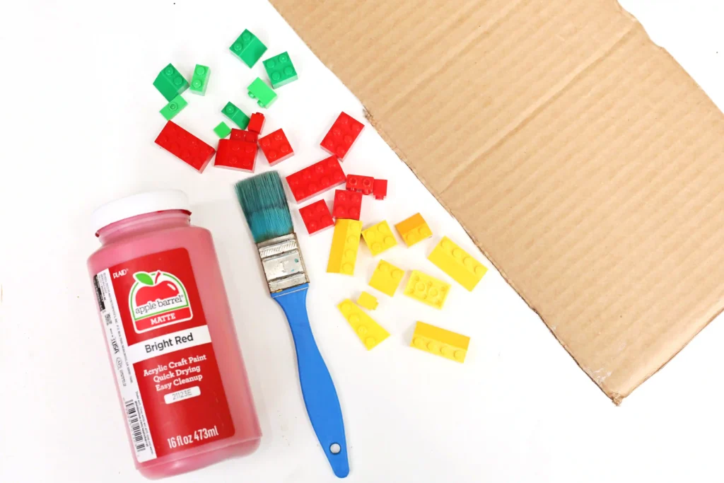 red acrylic paint bottle with a blue handle brush and scattered red, yellow, and green LEGO bricks on a white surface beside a cardboard corner.