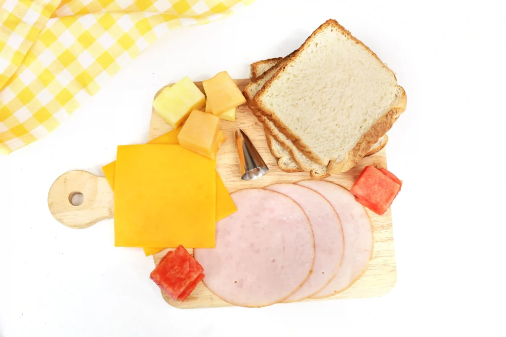 LEGO Lunchbox Idea - Cultivating Brilliant Minds ingredients for the lego lunch box idea : Assorted deli items on a wooden cutting board: slices of bread, ham, and cheese cubes with a yellow napkin nearby.