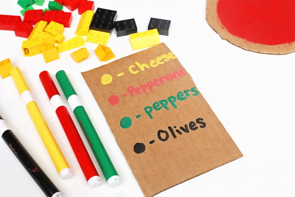 Crafting scene with colorful LEGO bricks and markers beside a cardboard pizza topping list (pepperoni, olives, etc.).