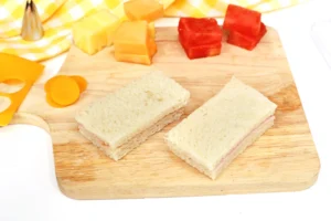Sandwich halves on a wooden cutting board with cheese cubes and orange slices in the background, ready to serve.