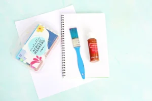 supplies for this letter o handprint craft: Blue paintbrush resting on a spiral notebook with an orange acrylic paint bottle and a pack of colorful markers on a white sheet of paper