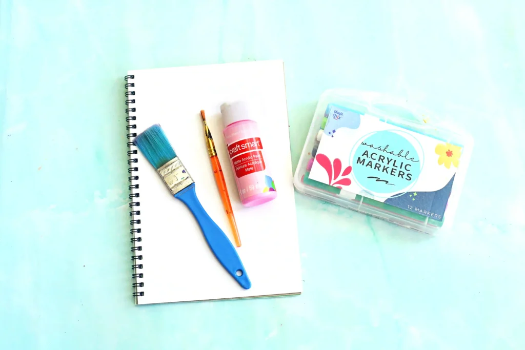 supplies for this pig craft for kids: Spiral notebook with paintbrush, pink acrylic paint bottle, pencil, and a marker set on a light blue surface.
