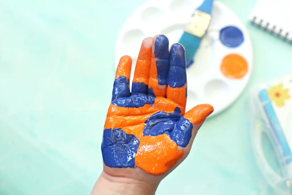 Hand raised with orange and blue paint smeared on fingers, backdrop of a painter's palette and art supplies.