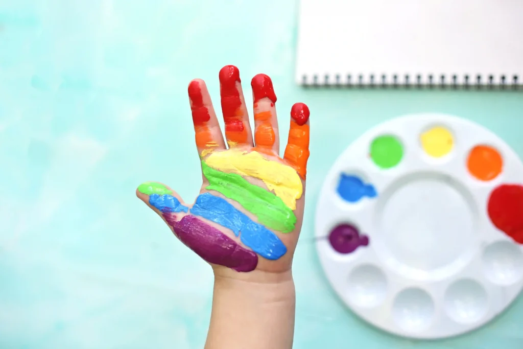 Raised hand covered in rainbow paint with a circular paint palette in the blurred background beside it. N/A