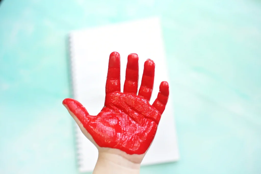 Red-painted hand raised in front of a pale, blurred background (notebook visible).abbreviated=