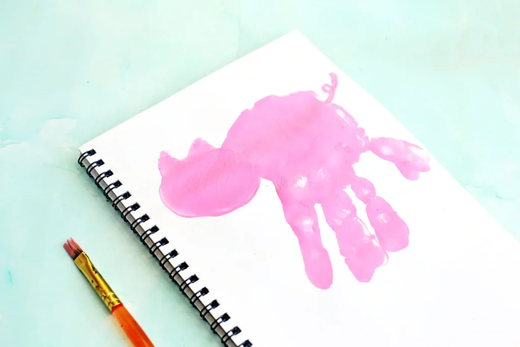 Spiral-bound notebook on a pale mint background with a pink paint handprint on the page with a pig head drawn on and a yellow paintbrush beside it.