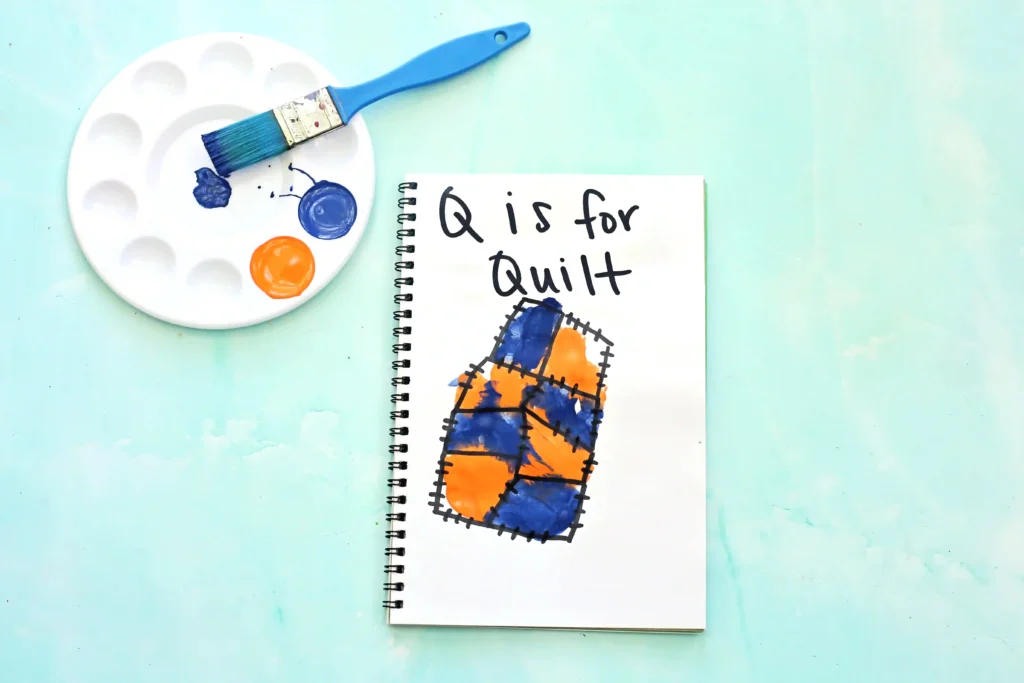 Notebook open to a page reading'Q is for Quilt' with a painted quilt block sketch, a blue paintbrush, and a white paint palette on a light blue background.