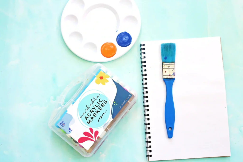 Artist's workspace with a blue brush, white notebook, paint palette (orange and blue) and paints in a transparent container on a mint background