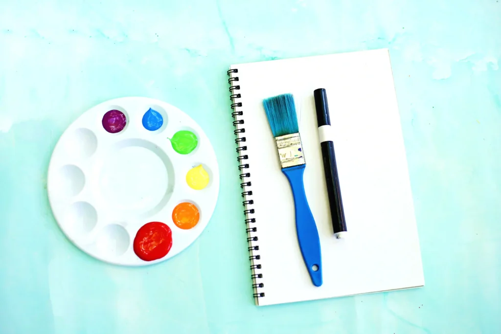 supplies for the letter r handprint craft: Flat lay of a white paint palette with bright paint blobs next to a spiral notebook, blue brush, and black marker on a light blue surface.