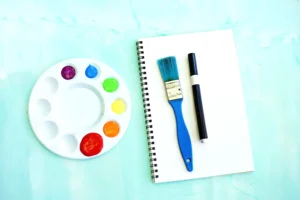 supplies for the letter r handprint craft: Flat lay of a white paint palette with bright paint blobs next to a spiral notebook, blue brush, and black marker on a light blue surface.
