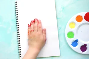 Child's hand with red and orange paint rests on a blank notebook, watercolor palette visible on the right side.