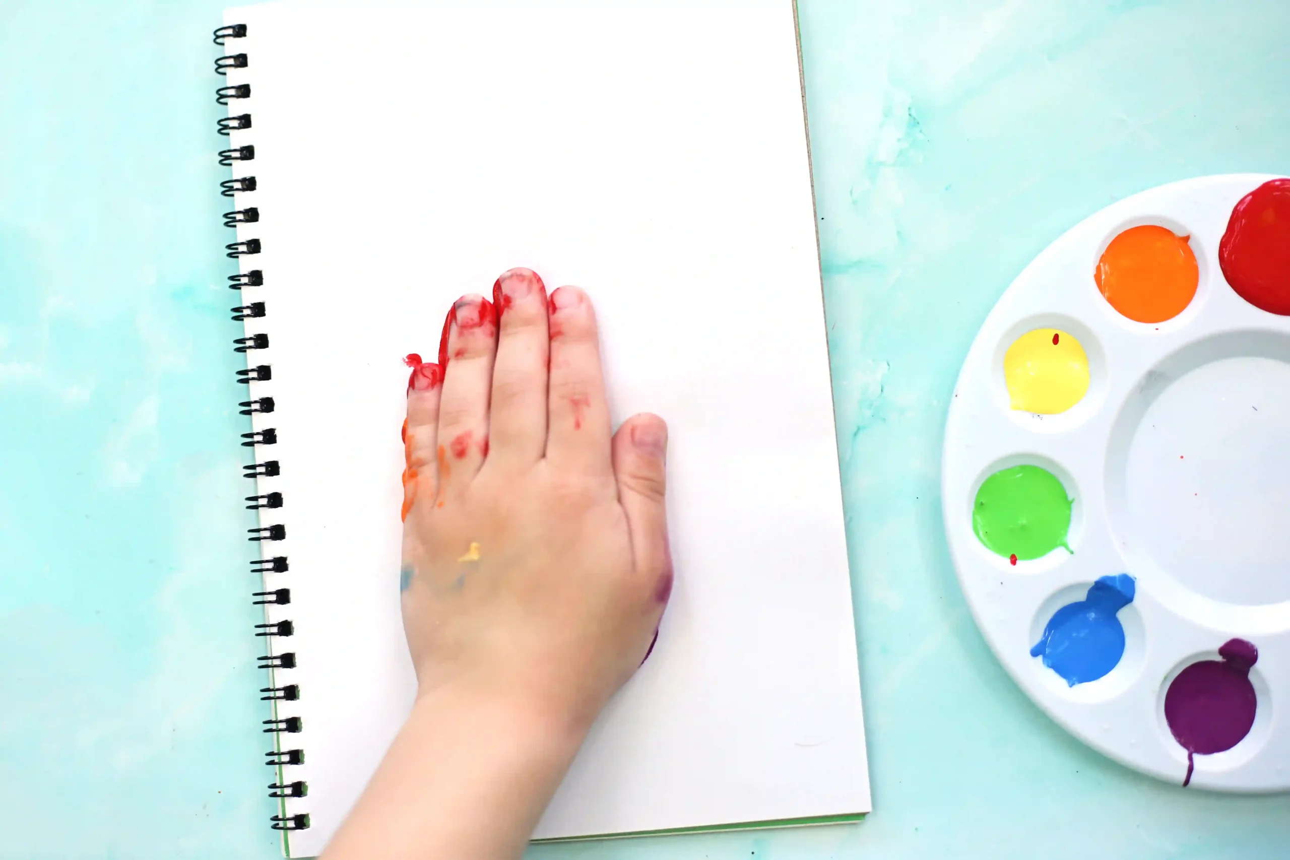 Child's hand with red and orange paint rests on a blank notebook, watercolor palette visible on the right side.