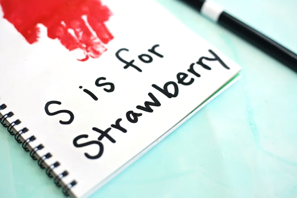 Close-up of a spiral notebook page reading'S is for Strawberry' in black handwriting, with a red paint blot and a black pen on a light blue surface.