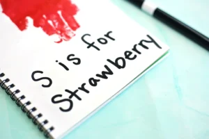 Close-up of a spiral notebook page reading 'S is for Strawberry' in black handwriting, with a red paint blot and a black pen on a light blue surface.