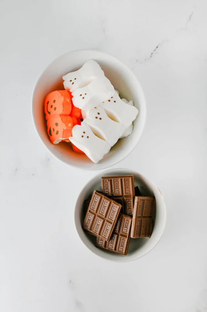ingredients for the halloween d'mores dip: Bowl of Halloween marshmallows—white ghost shapes and orange pumpkin shapes—stacked above a bowl of Hershey's chocolate bars on a light marble surface.