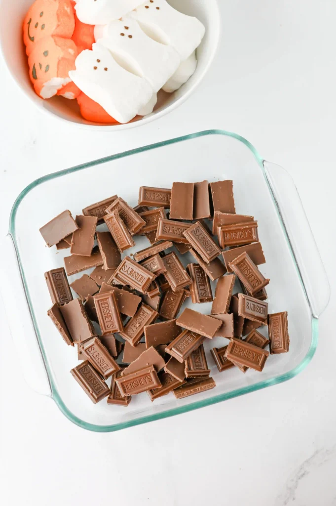 Milk chocolate pieces with Hershey's embossed, in a glass dish next to a bowl of orange and white marshmallows.