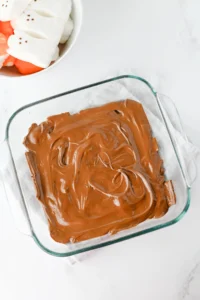 Glass baking dish filled with melted milk chocolate on a white surface, marshmallows visible in a separate bowl nearby.