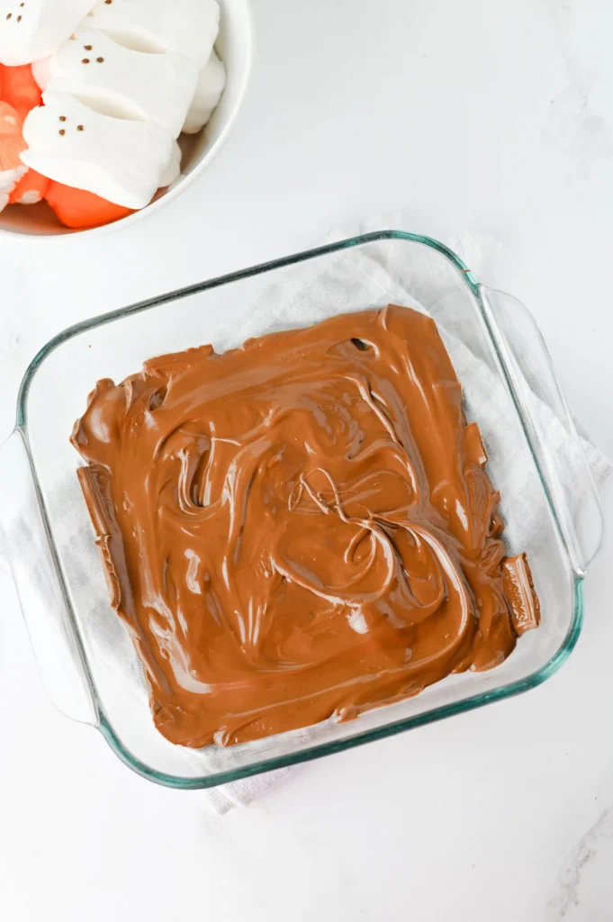 Glass baking dish filled with melted milk chocolate on a white surface, marshmallows visible in a separate bowl nearby.