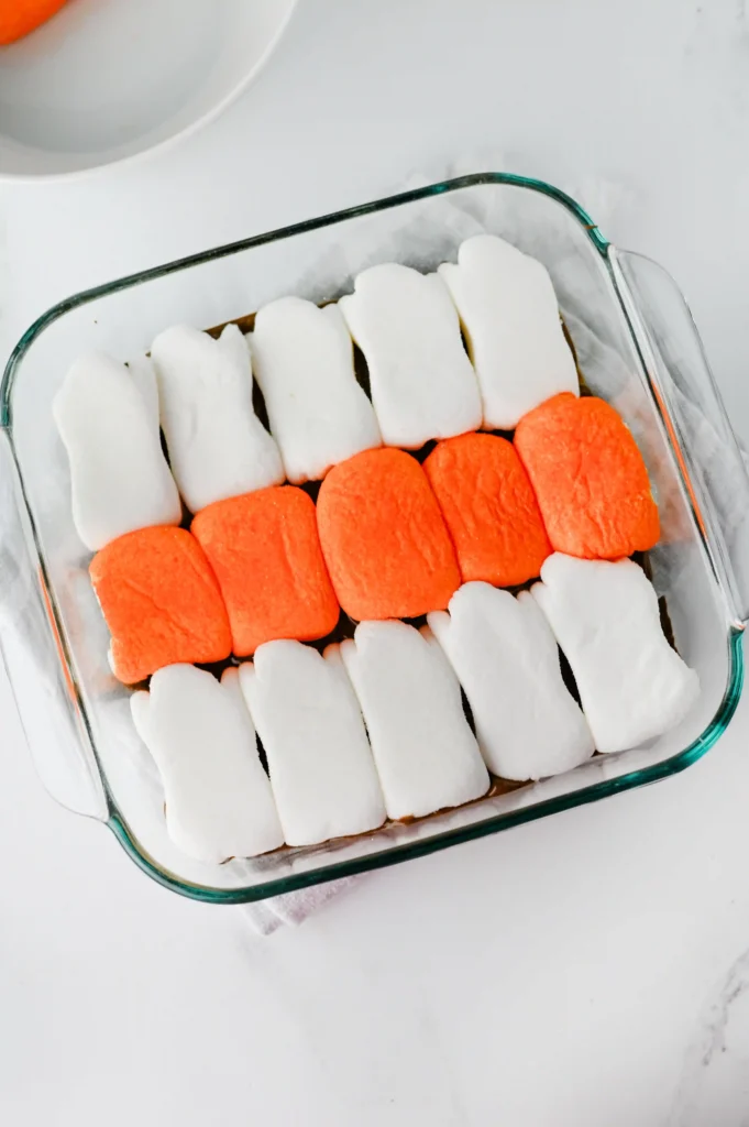 Glass baking dish with alternating rows of orange and white marshmallows over a chocolate base in a pan.