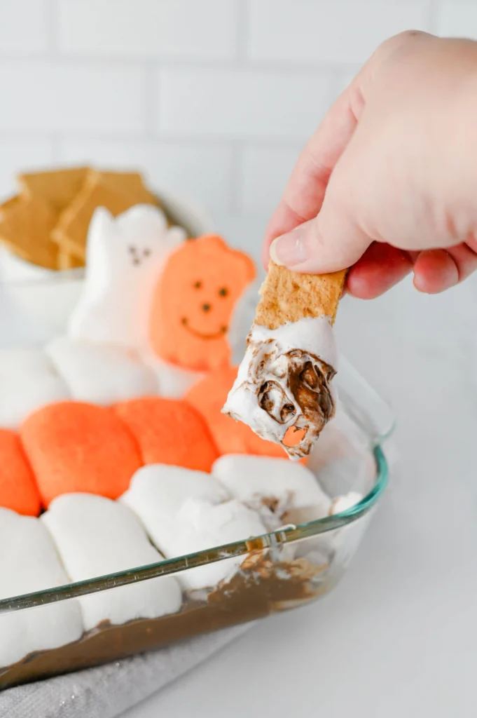Hand dipping a biscuit into melted chocolate over a tray of marshmallows and Halloween cookies in the background with ghost and pumpkin shapes