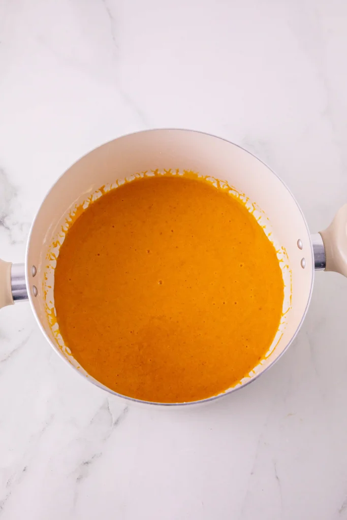 Top-down view of a white saucepan filled with bright orange batter on a marble countertop.