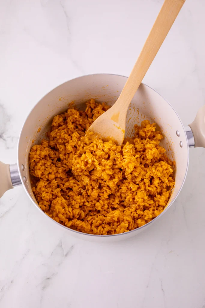 Pot of orange cooked rice krispies cereal with a wooden spoon resting inside the pan.