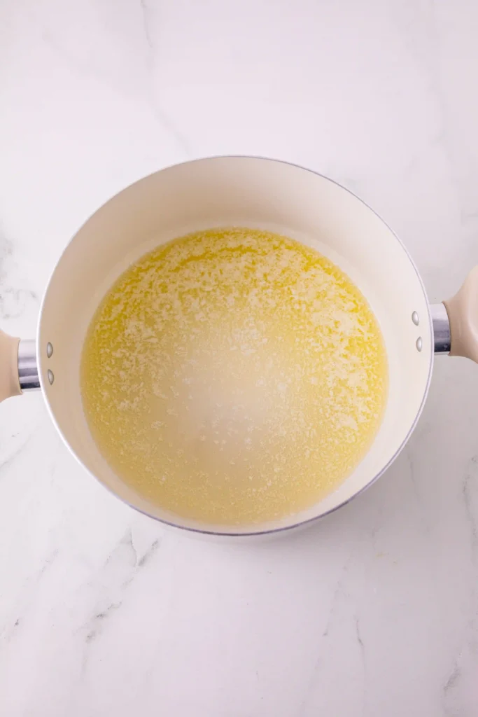 Top-down view of a white saucepan on a marble surface, filled with pale yellow melted butter.