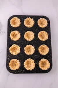 Nonstick muffin pan with twelve cups filled with rice krispie cereal-based batter on a light marble surface.
