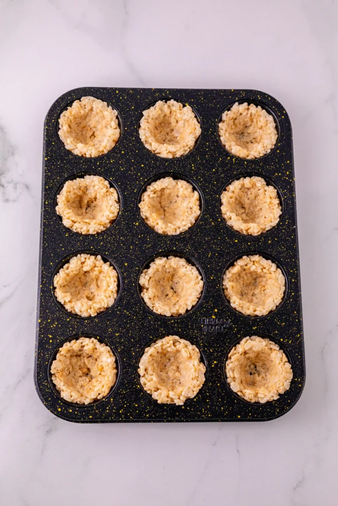 Nonstick muffin pan with twelve cups filled with rice krispie cereal-based batter on a light marble surface.