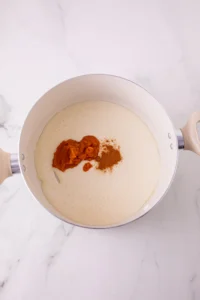 Top-down view of a white saucepan with batter and a dollop of orange pumpkin puree plus brown spice on top, on a marble counter.