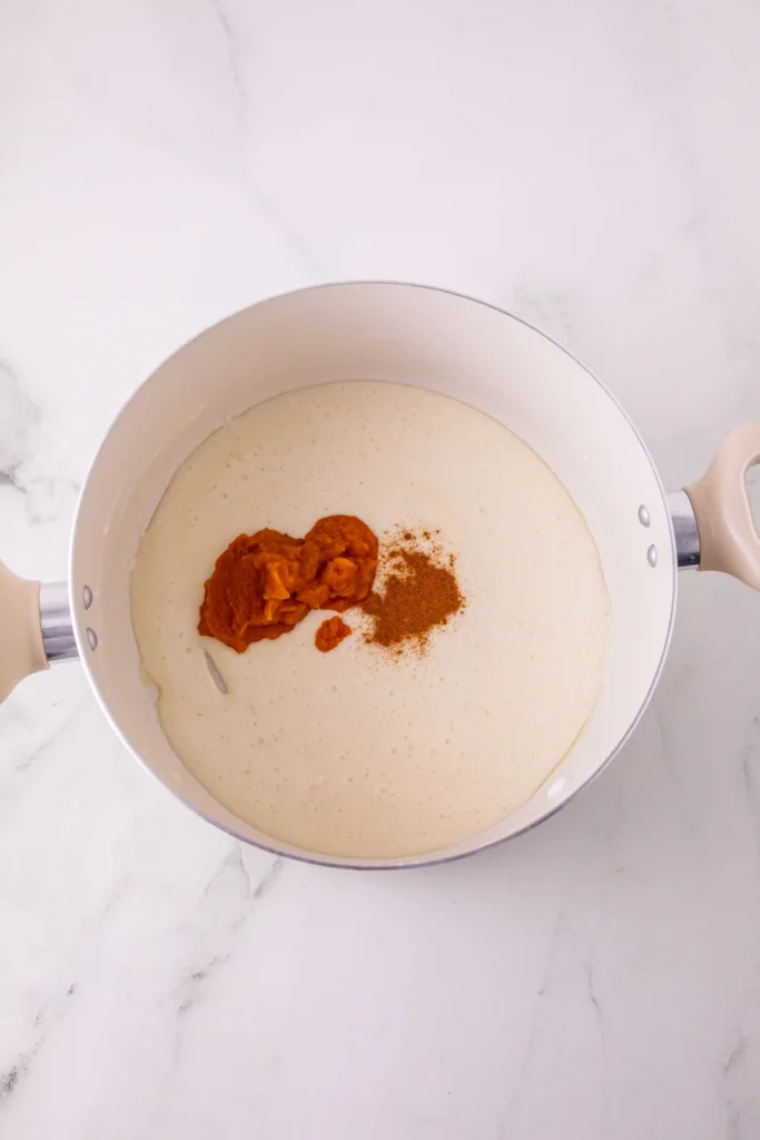 Top-down view of a white saucepan with batter and a dollop of orange pumpkin puree plus brown spice on top, on a marble counter.