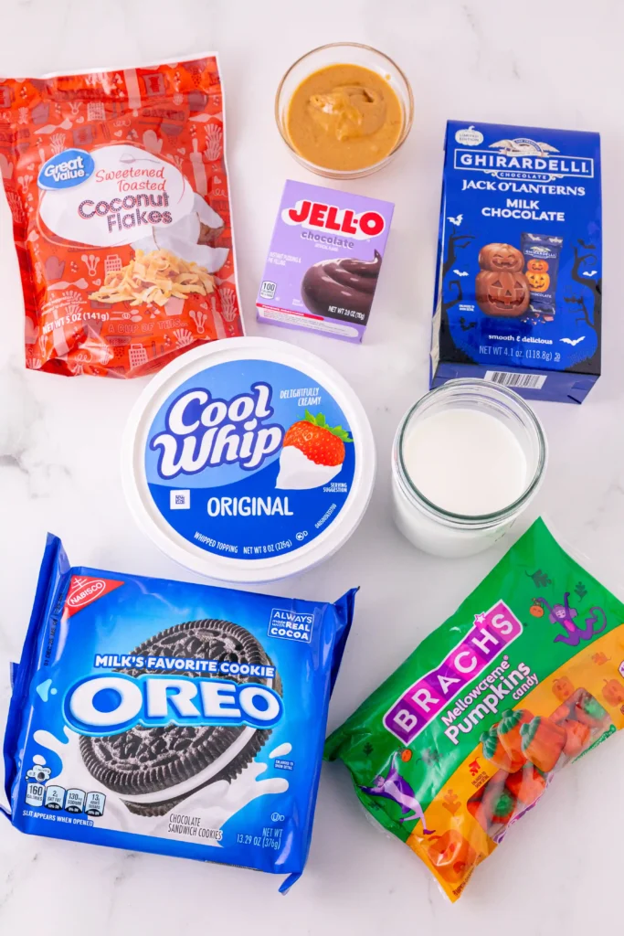 ingredients for the pumpkin patch dirt cups: coconut flakes, Jell-O chocolate mix, Ghirardelli milk chocolate, Cool Whip, Oreo cookies, Brach's pumpkin candy, a bowl of peanut butter and a glass of milk.