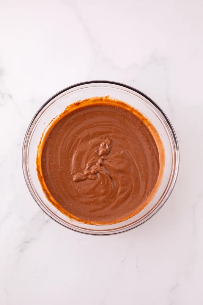 Top-down view of a glass bowl filled with glossy chocolate pudding mix on a white marble surface.