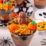 Halloween-themed dessert cups topped with chocolate pumpkin figures, orange candy pumpkins, and orange sprinkles over cookie crumble.