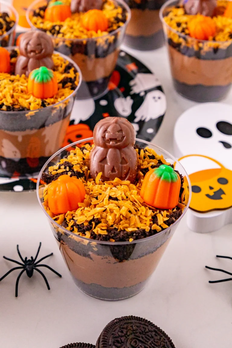 Halloween-themed dessert cups topped with chocolate pumpkin figures, orange candy pumpkins, and orange sprinkles over cookie crumble.
