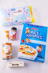 ingredients for the pumpkin pie rice krispies: Rice Krispies cereal, mini marshmallows, pumpkin spice, canned pumpkin, butter, and a squeeze tube on a white surface.