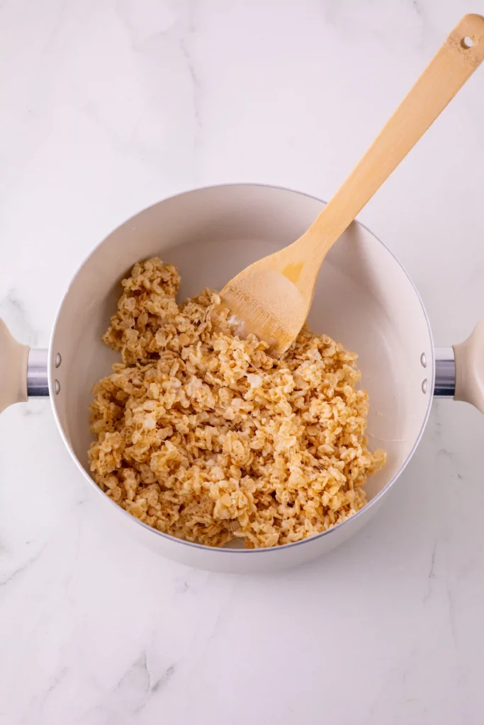 Pot in use with a wooden spoon stirring a large amount of rice krispies cereal inside a stainless pot on a marble surface.