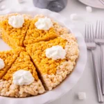 Rice crispy pumpkin pie treats cut into triangles on a white plate, topped with dollops of whipped cream or icing sugar.
