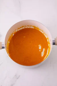 Overhead view of a white saucepan filled with orange pumpkin pie puree on a light marble countertop.