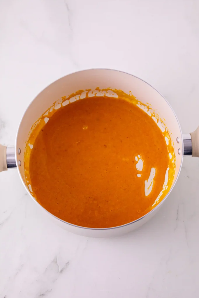 Overhead view of a white saucepan filled with orange pumpkin pie puree on a light marble countertop.