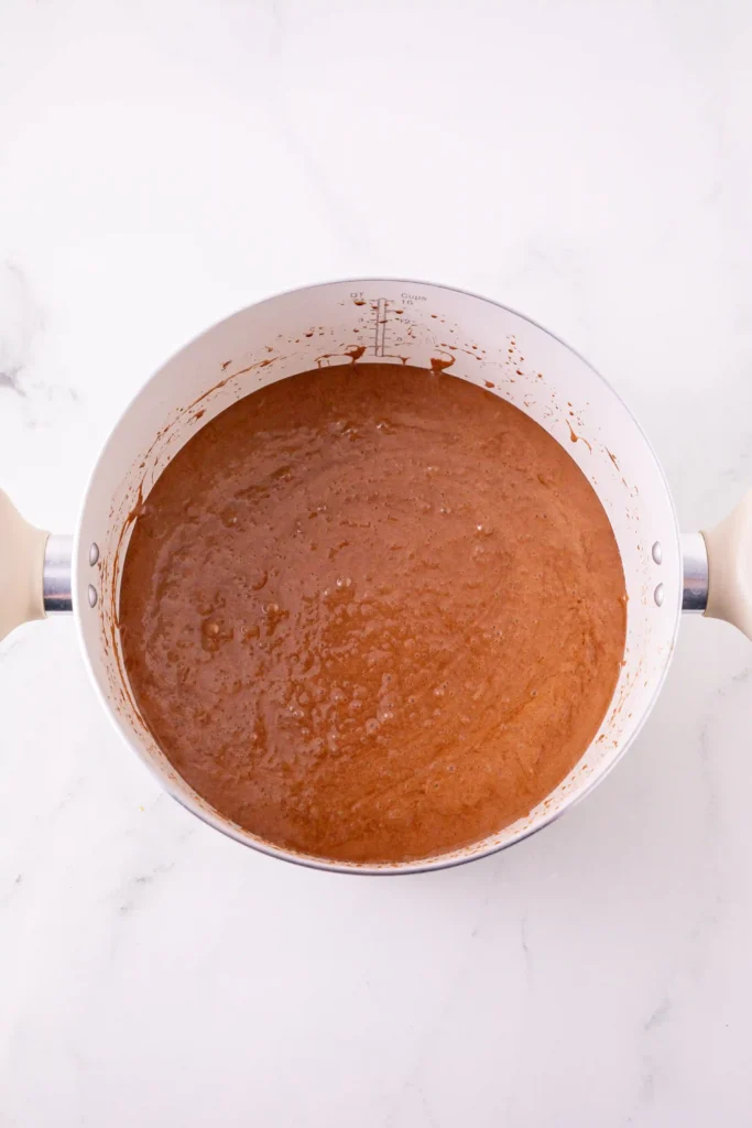 Overhead view of a white saucepan filled with brown batter, with batter splatters along the rim on a white marble surface.